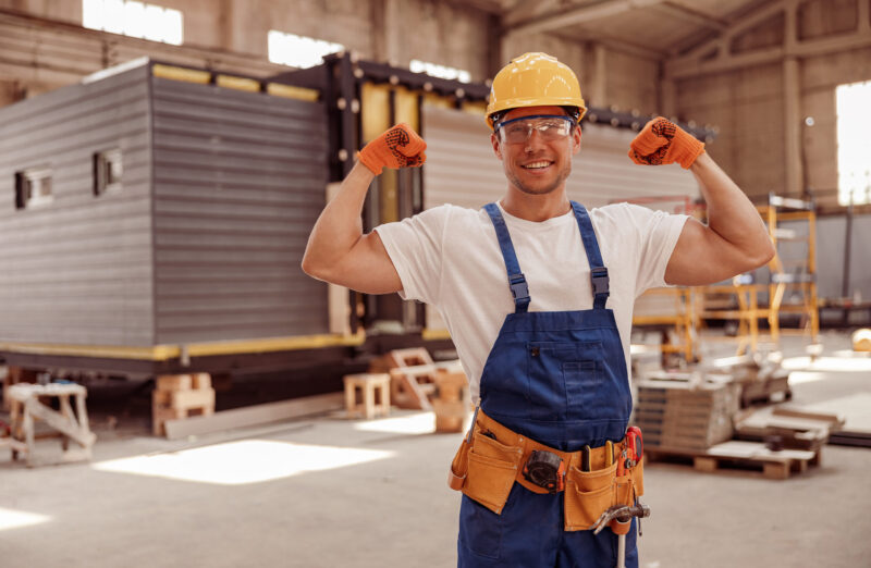 Handsome athletic man construction worker looking at camera and smiling while showing perfect biceps muscle